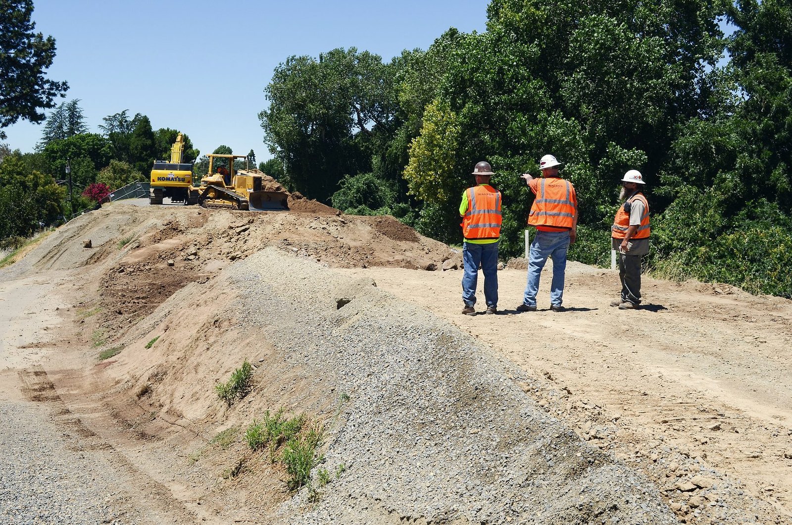 Three crew members in hard hats and orange vests on a levee embankment while a dozer works behind them